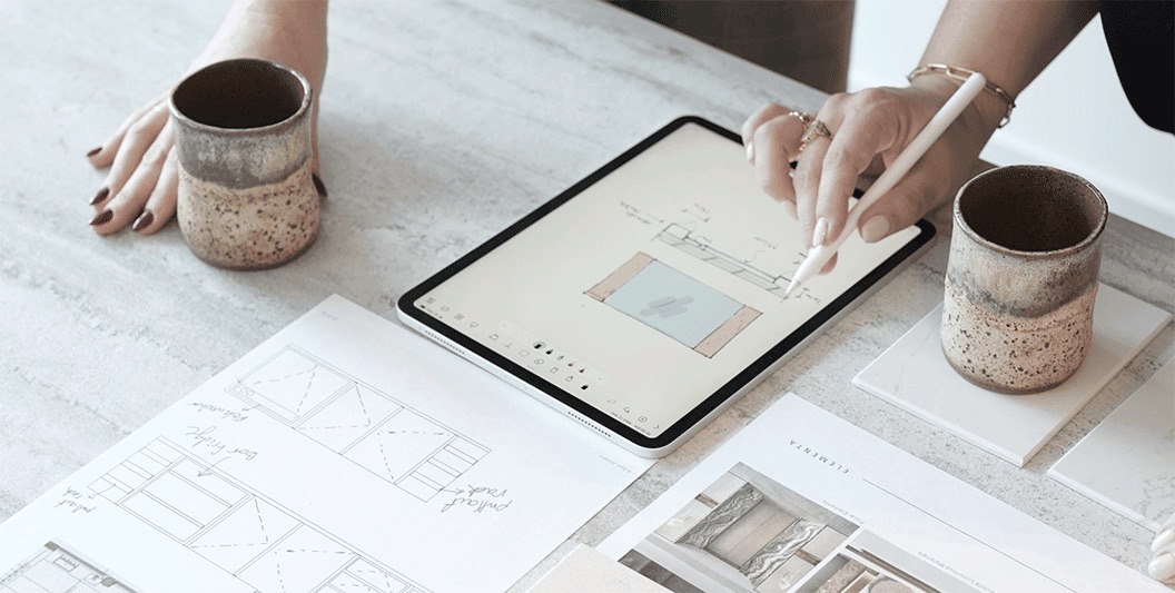 A designer sketching a room layout on a tablet beside floor-plan drawings and two speckled ceramic cups on a marble table